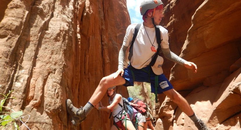 A person wearing safety gear braces their feet against the walls of a narrow canyon. Others are behind them.